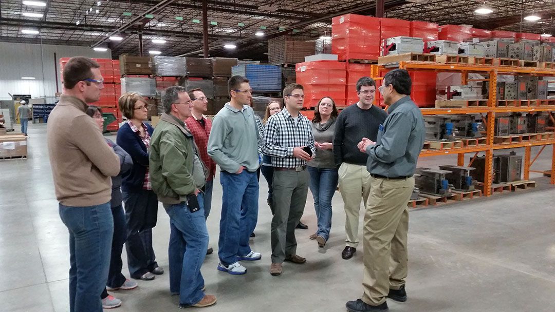 A group of people listens to a speaker in a large warehouse with orange storage racks filled with boxes.
