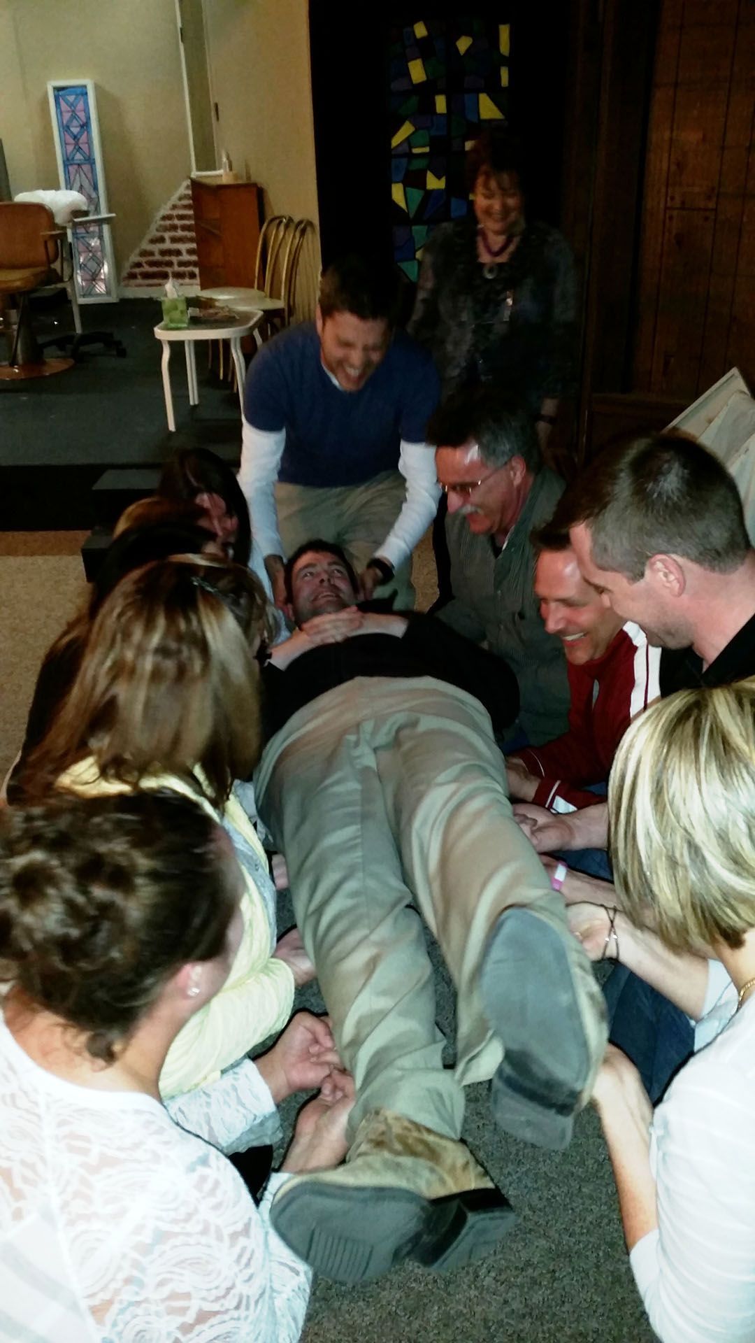 A group of people gathers around a person lying on the floor, holding their feet in a workshop setting.