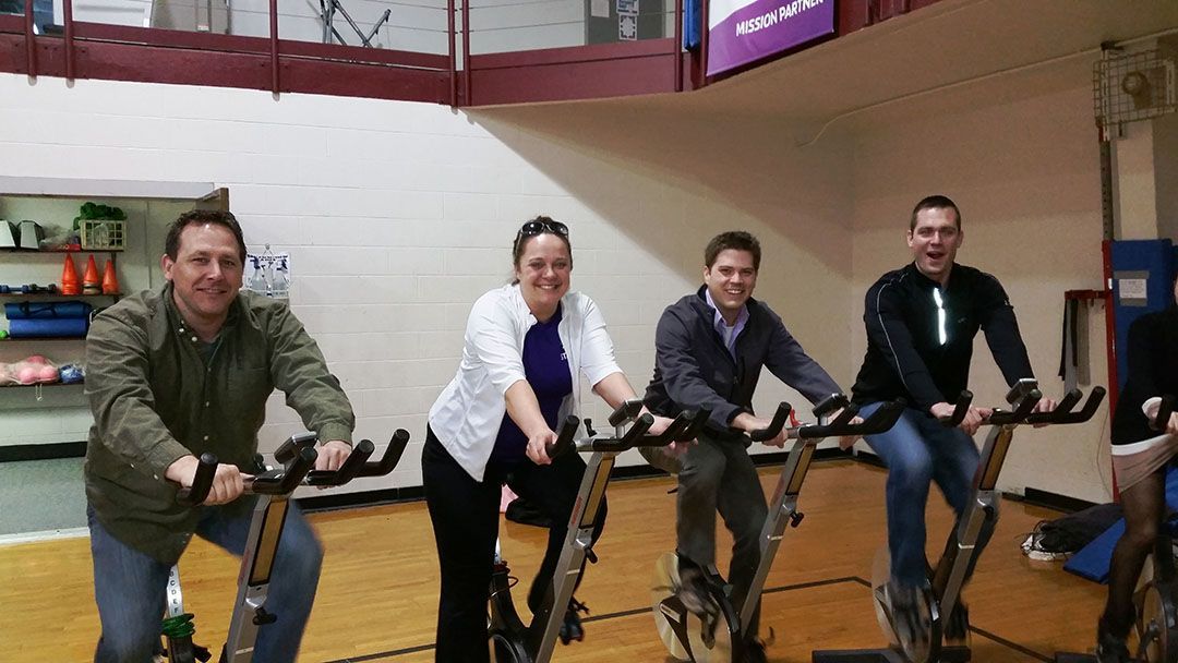 Four people smile while sitting on stationary exercise bikes in a gym.