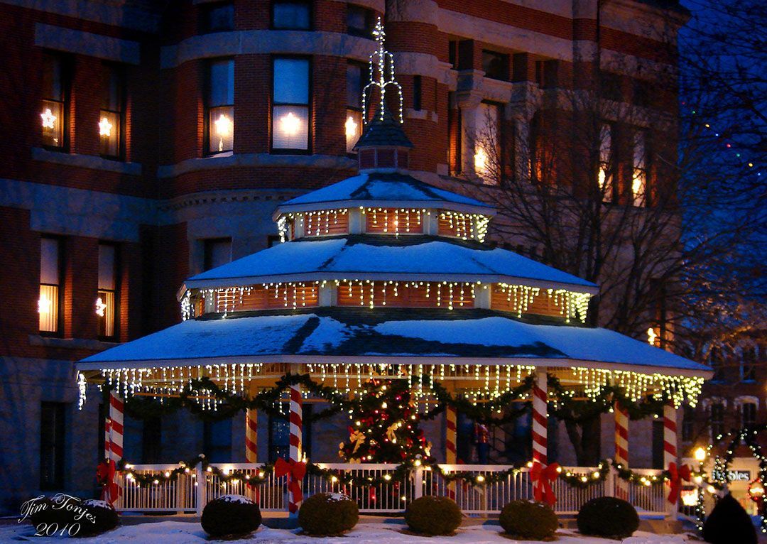 A snow-covered gazebo decorated with Christmas lights and candy-cane-striped pillars in front of a building at night.