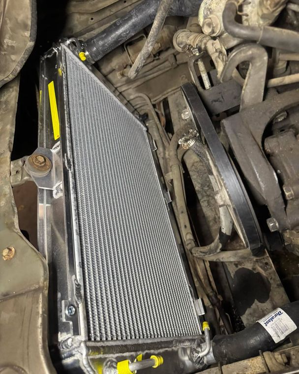 Car engine bay with a silver aftermarket radiator installed beside hoses and belts.