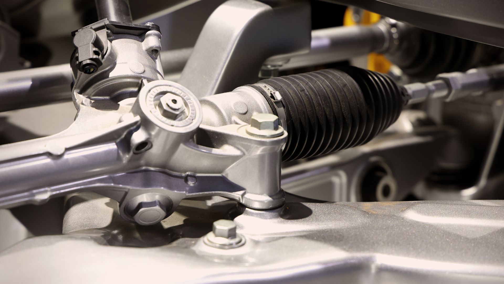 Close-up of a metallic steering rack and suspension components with black rubber boot under a car hood