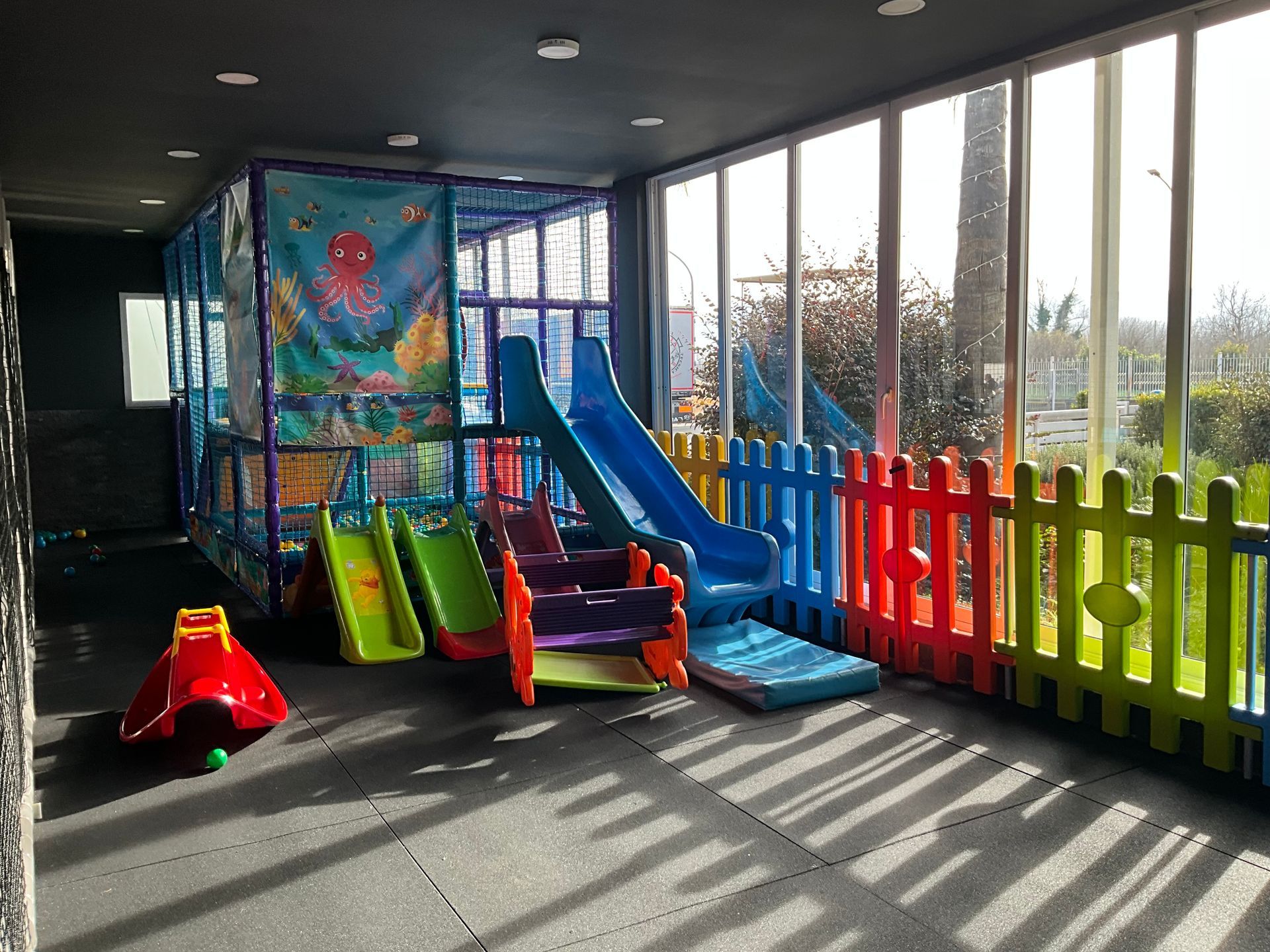 Area giochi interna per bambini con scivoli, una struttura per arrampicarsi e una recinzione colorata vicino alle finestre.