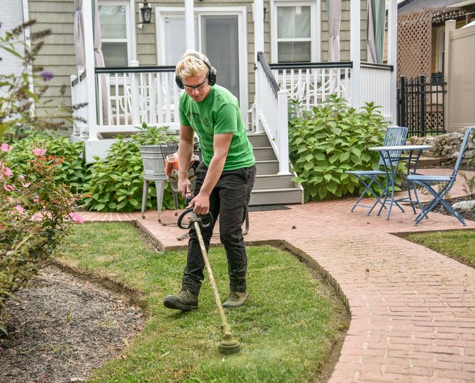 Person mowing a small front lawn beside a brick path, with a house porch and patio chairs in the background