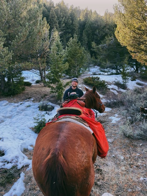 Man riding a brown horse on a snowy trail in a forest. Horse's rear is in focus, red blanket visible.