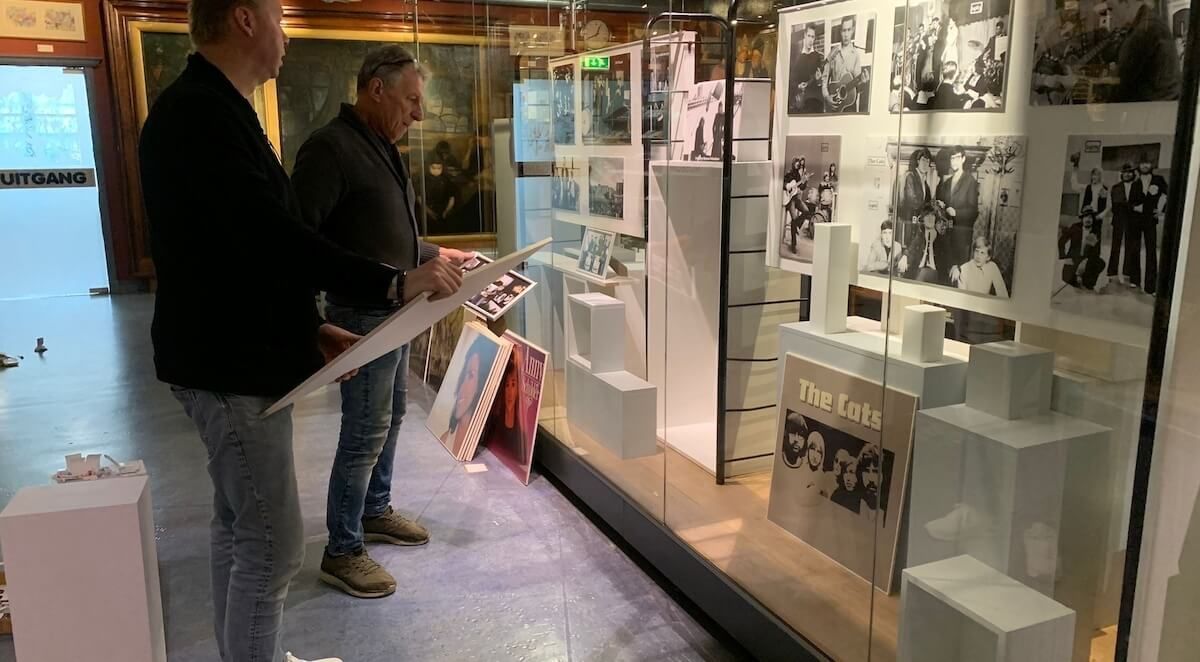 Twee mannen bekijken een tentoonstelling in een museum. Vitrines met foto's en objecten zijn zichtbaar.