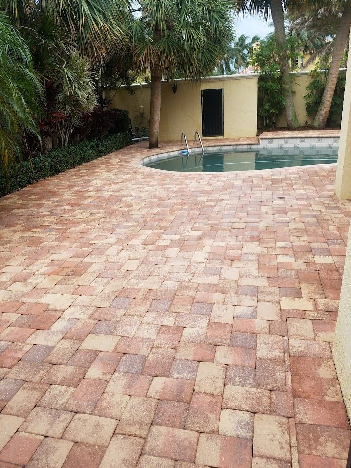 Brick-paved patio beside a swimming pool, bordered by lush tropical plants and a small pool house