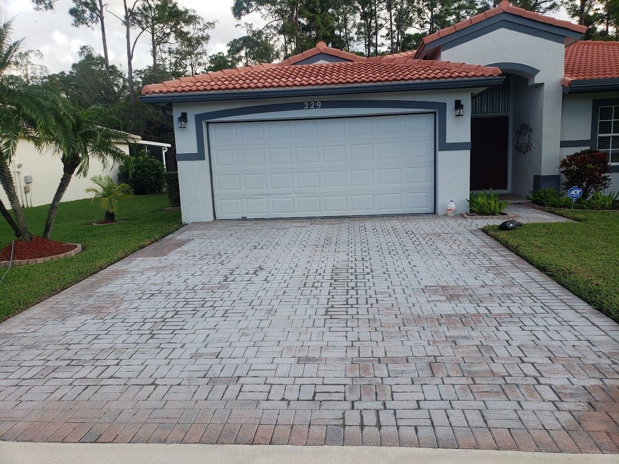 Suburban house with white garage door and brick driveway, bordered by grass and trees