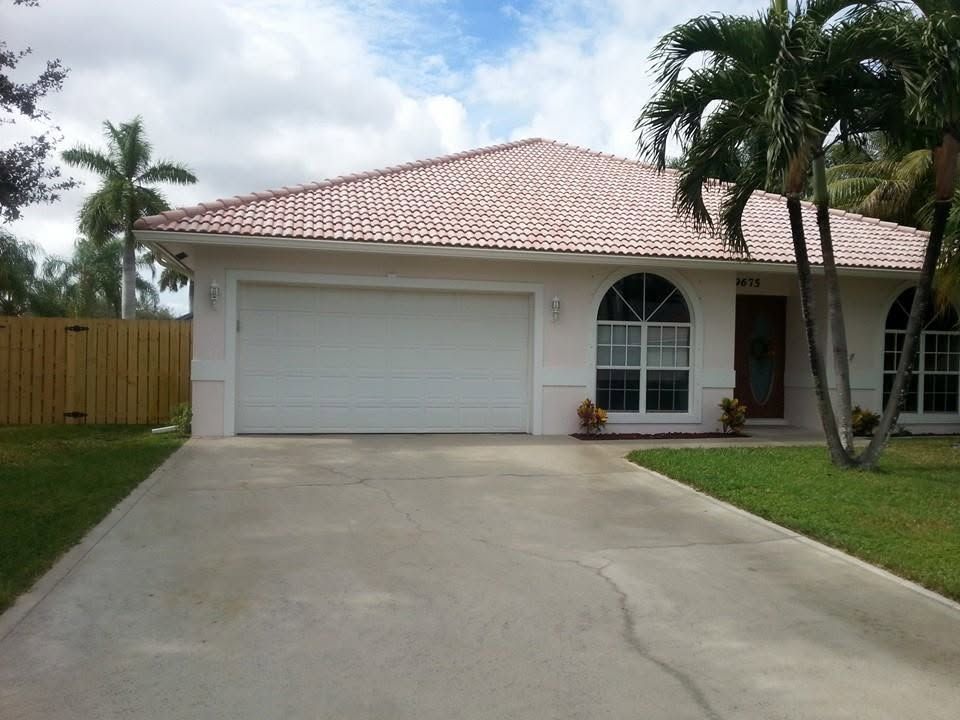 Single-story suburban house with a white garage, tiled roof, and driveway lined by palm trees.