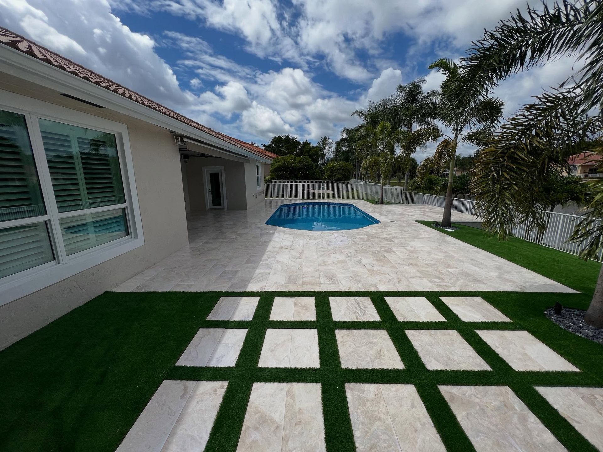 Backyard patio with pool, white tile, palm trees, and green grass under a cloudy sky