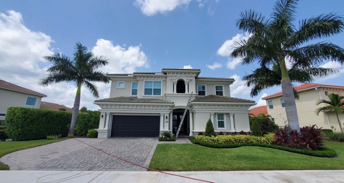 Single-story suburban house with a white garage, tiled roof, and driveway lined by palm trees.