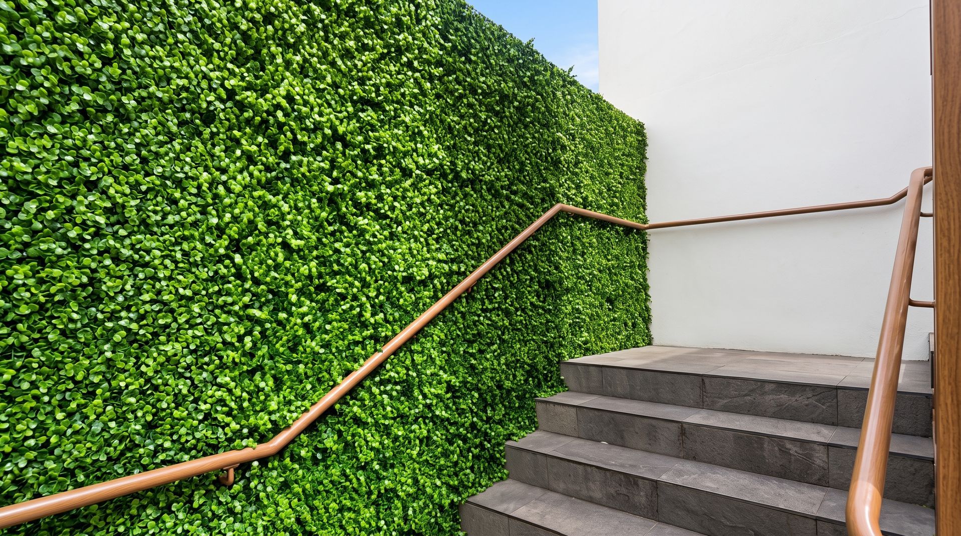 Staircase with gray steps and wooden handrails beside a bright green hedge wall