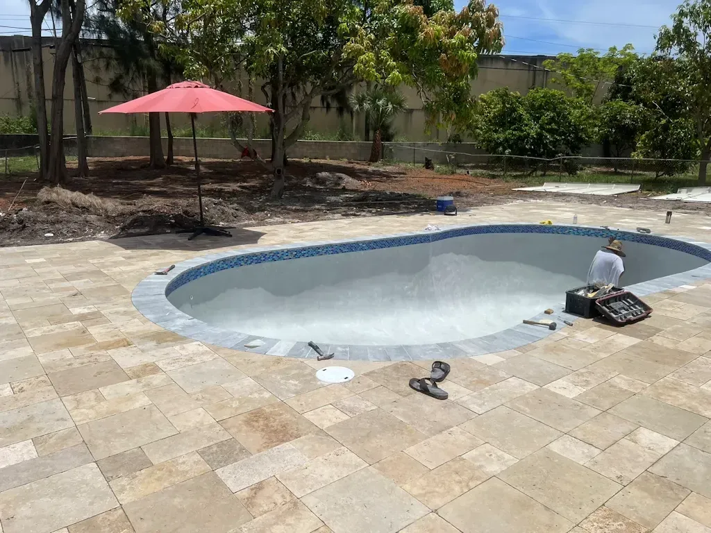 Empty backyard pool being resurfaced, with stone patio, red umbrella, and trees in the background.