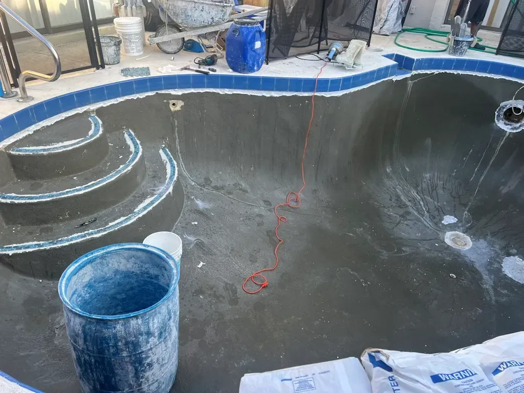 Empty pool being resurfaced, with dark coating, blue edge trim, buckets, and tools nearby
