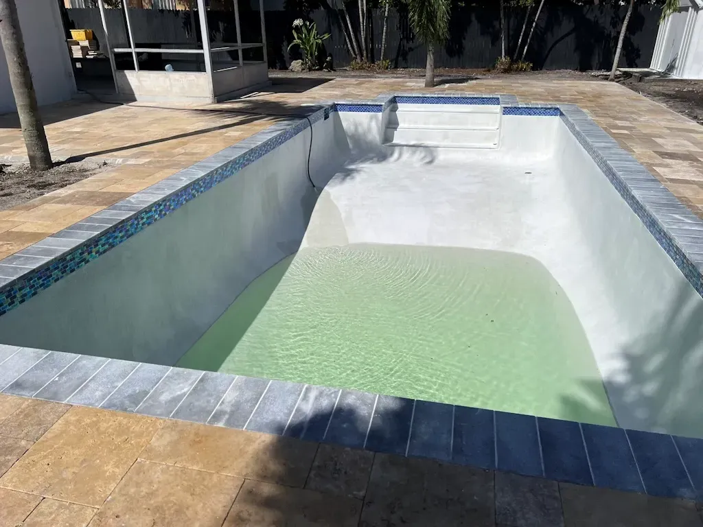 Empty drained swimming pool with a pale green patch of water at one end, surrounded by tiled coping.
