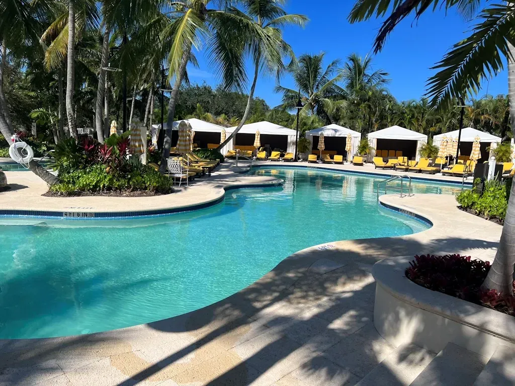 Resort swimming pool with turquoise water, palm trees, and white cabanas under a bright blue sky