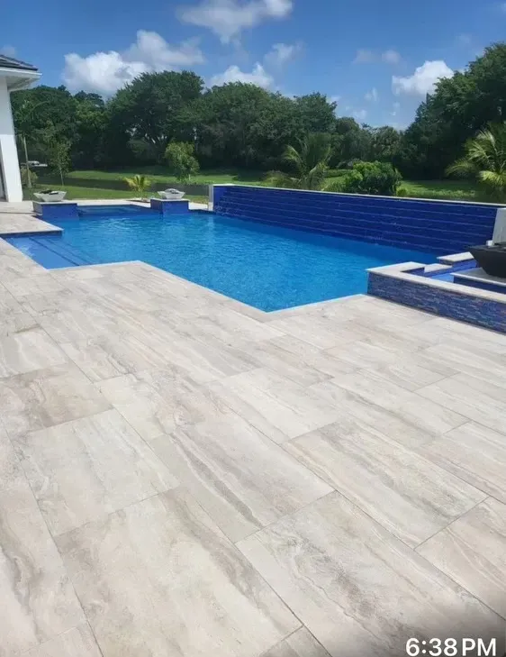 Backyard pool with blue water, light stone patio, and green trees under a sunny sky