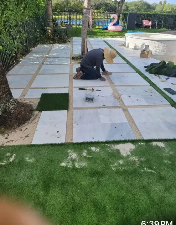 Worker laying pavers on a backyard patio beside a lawn and pool area