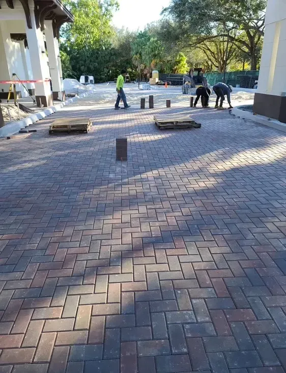 Workers laying brick pavers on a courtyard walkway beside a building, with trees in the background
