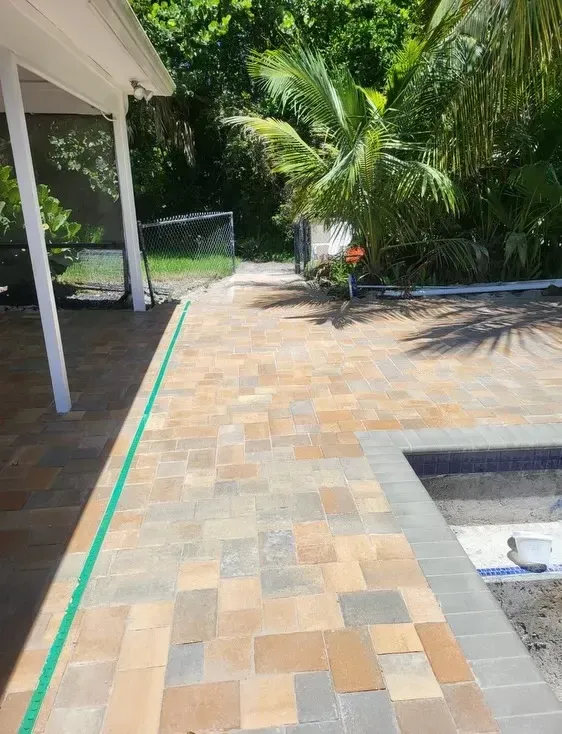 Tiled patio beside a pool, with palm trees, a screened area, and green border along the walkway.