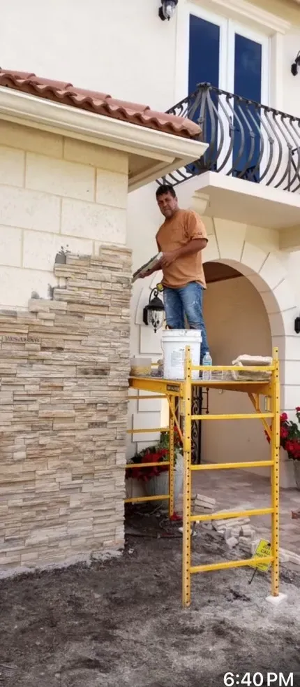 Worker on a yellow scaffold painting a house exterior with stone and white walls.