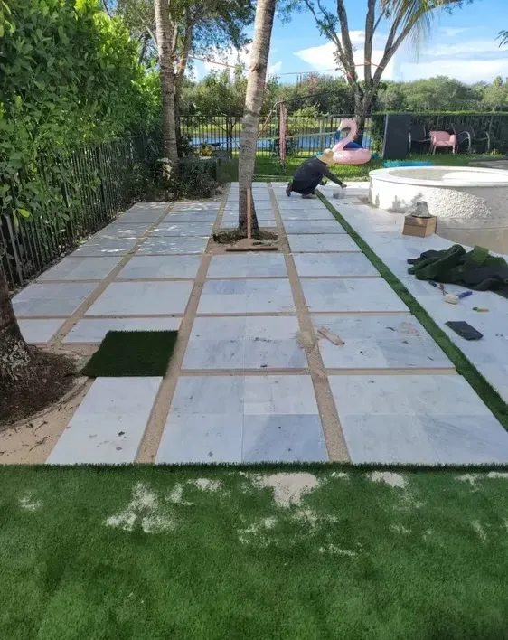 Backyard patio under construction with white pavers, wooden spacers, and a worker near a raised spa