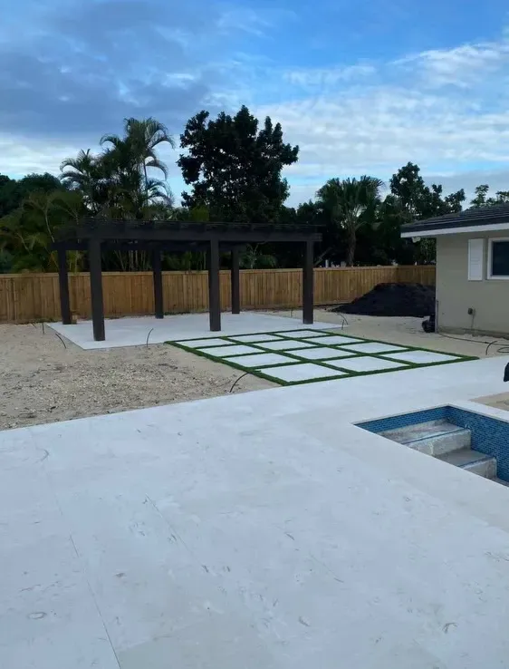 Backyard patio with pergola, stepping stones, pool edge, and fenced yard under a partly cloudy sky