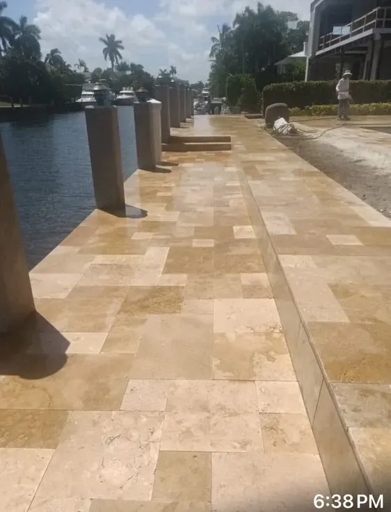 Sunlit tiled waterfront walkway beside water, with railings and a person in the distance