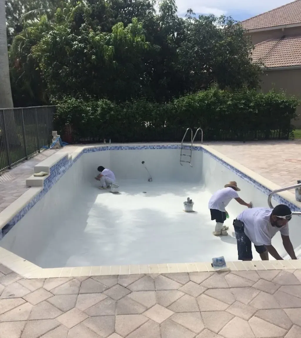 Two people cleaning an empty white backyard swimming pool beside a patio and hedge.