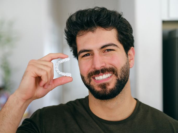 Smiling man holds a clear dental aligner beside his face in a bright room