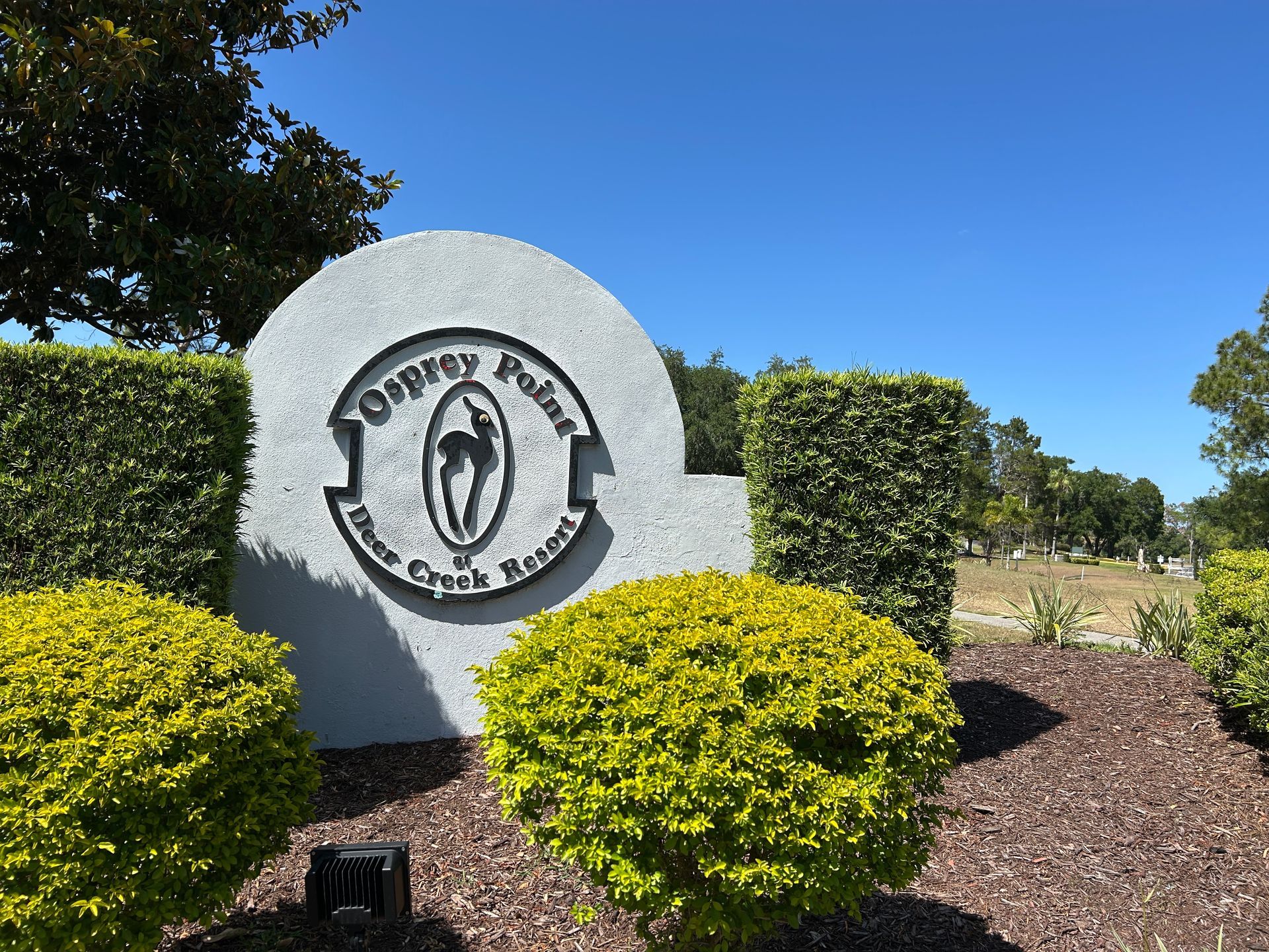 Community entrance sign on a white stone monument, surrounded by trimmed bushes under a blue sky