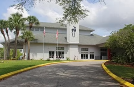 Modern white building with flags, trees, and a curved driveway under cloudy skies