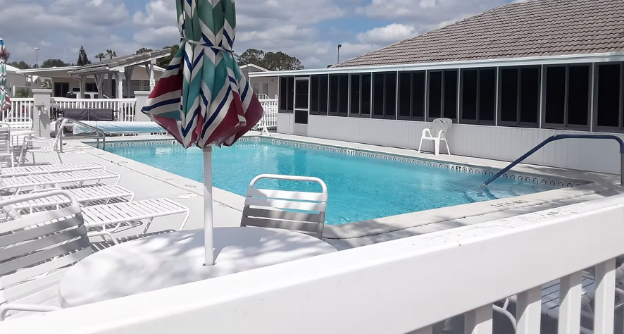 Outdoor swimming pool with white lounge chairs and a striped umbrella beside a building with screened windows