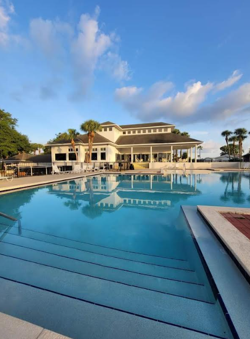 Modern resort pool with clear blue water, lounge chairs, and a white clubhouse under a partly cloudy sky