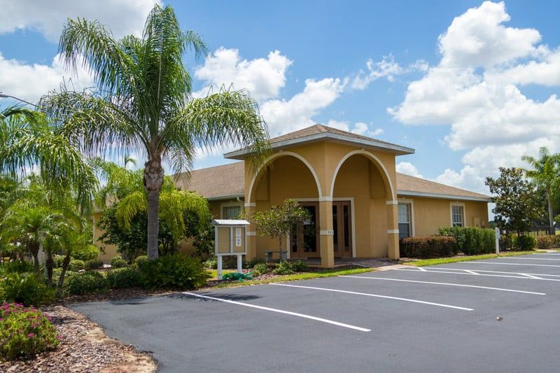 Tan stucco building with arched entrance and palm trees beside an empty parking lot under a blue sky