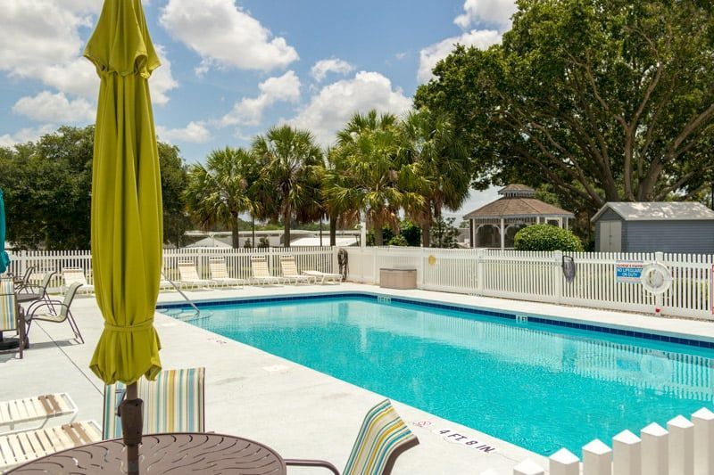 Outdoor swimming pool with blue water, white fence, green umbrella, and palm trees under a partly cloudy sky