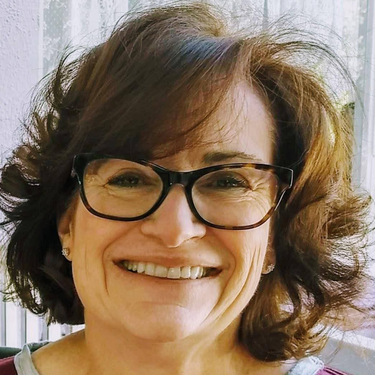 Smiling woman with black glasses and wavy brown hair in front of a bright curtain