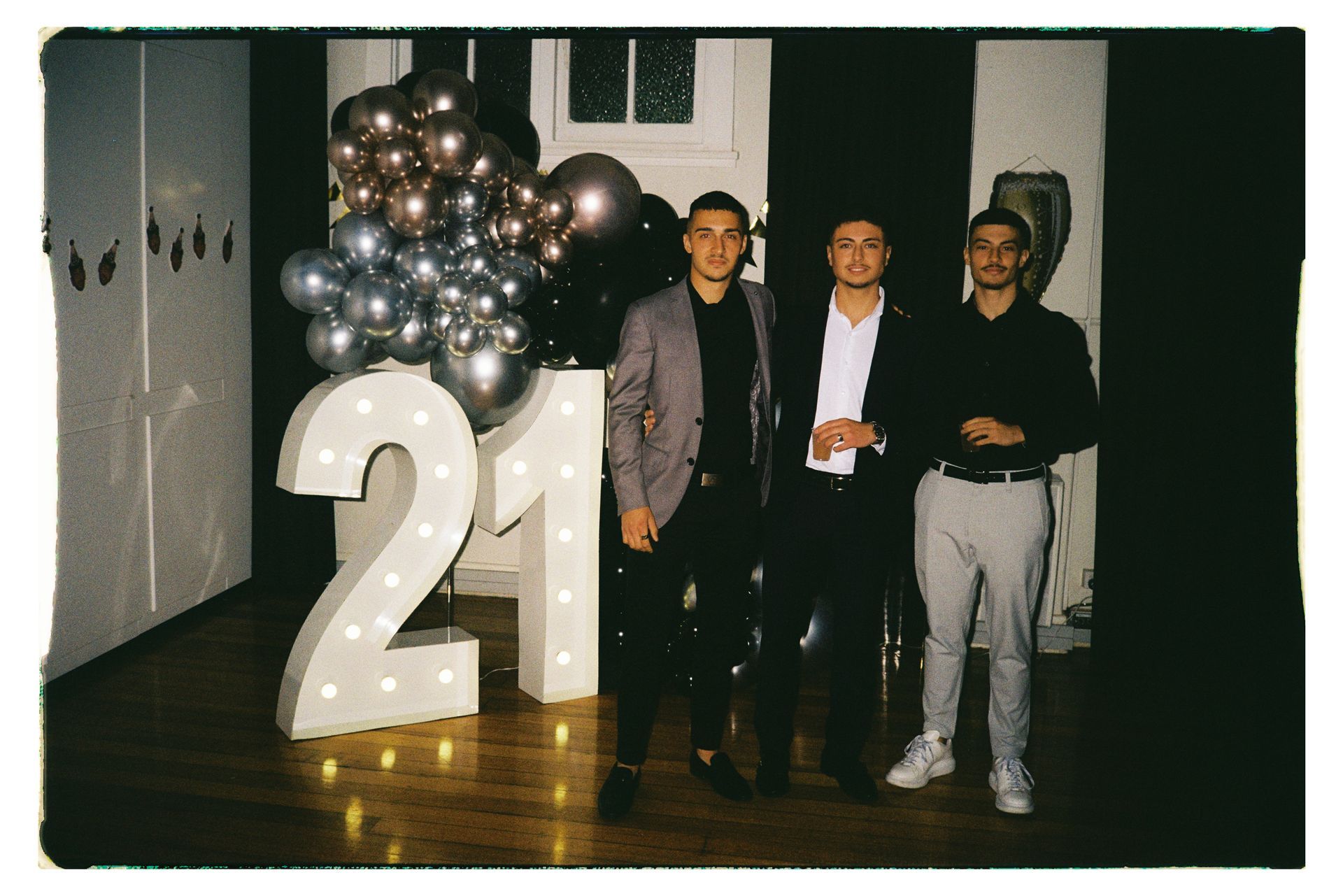 Three people posing beside a large “21” and silver balloon display at a party.