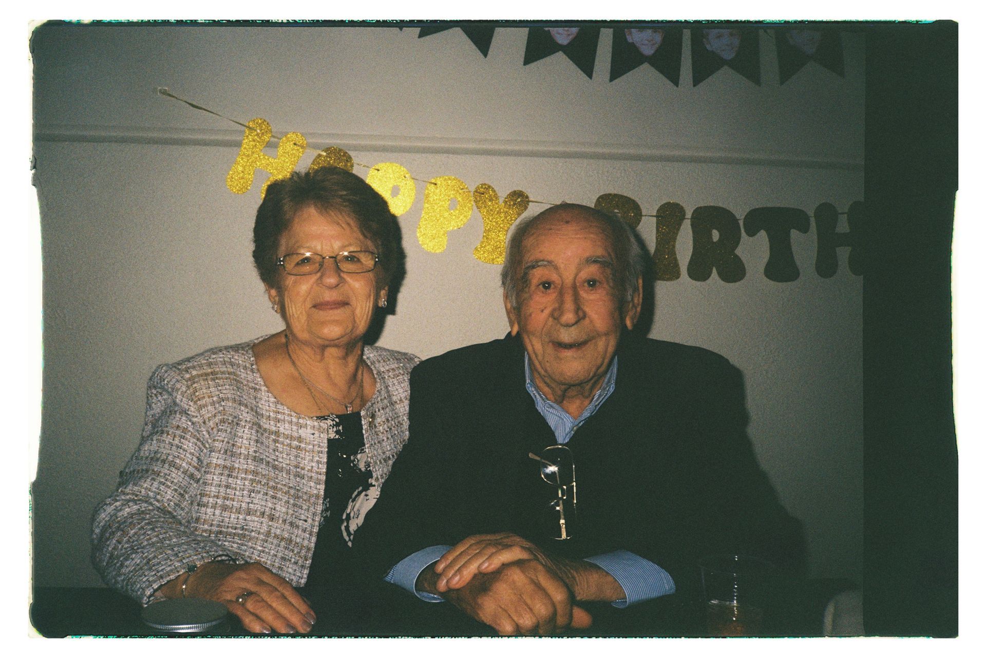 Two older adults seated at a party table, smiling in front of a “Happy Birthday” banner.