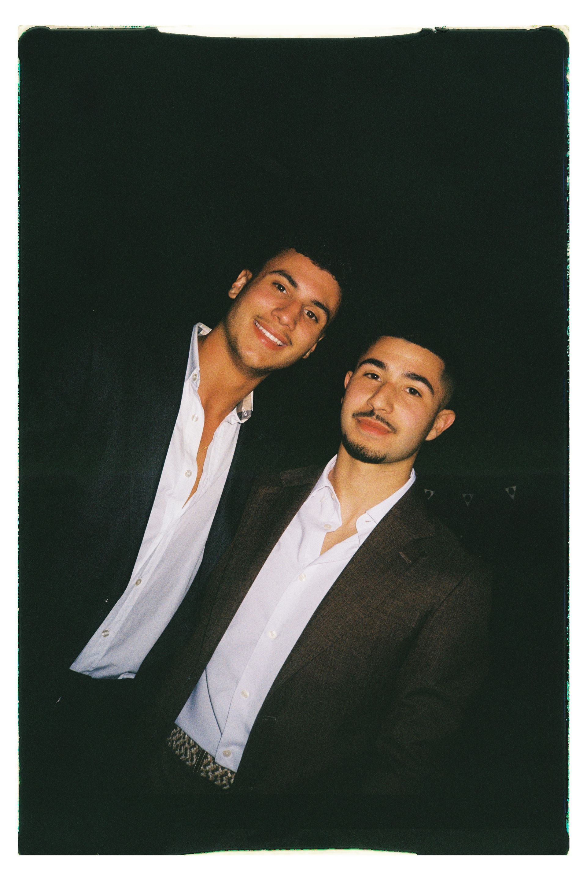 Two smiling men in white shirts and dark jackets posing against a dark background