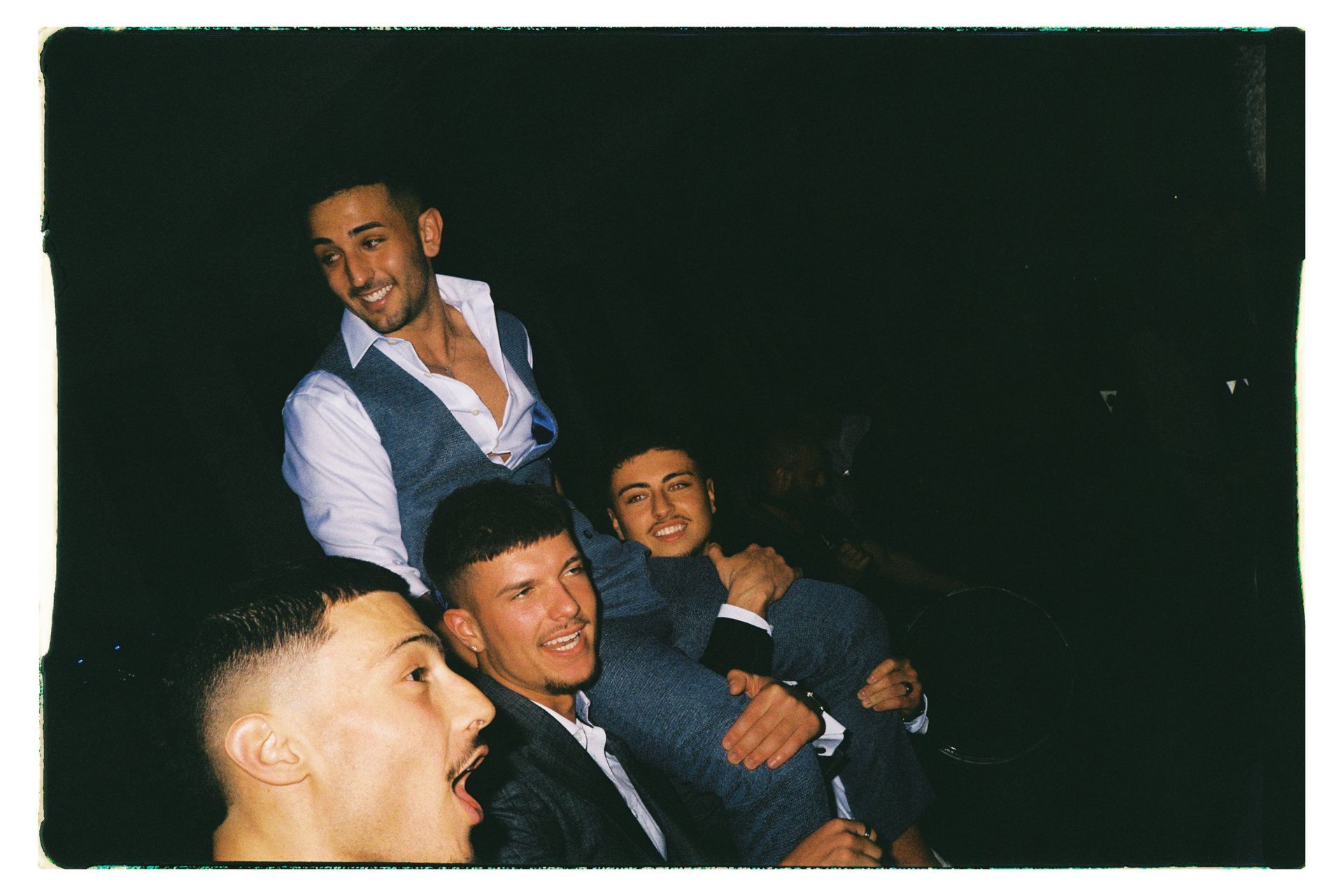 Four people smiling and posing closely together in a dark setting, one in a white shirt and vest.