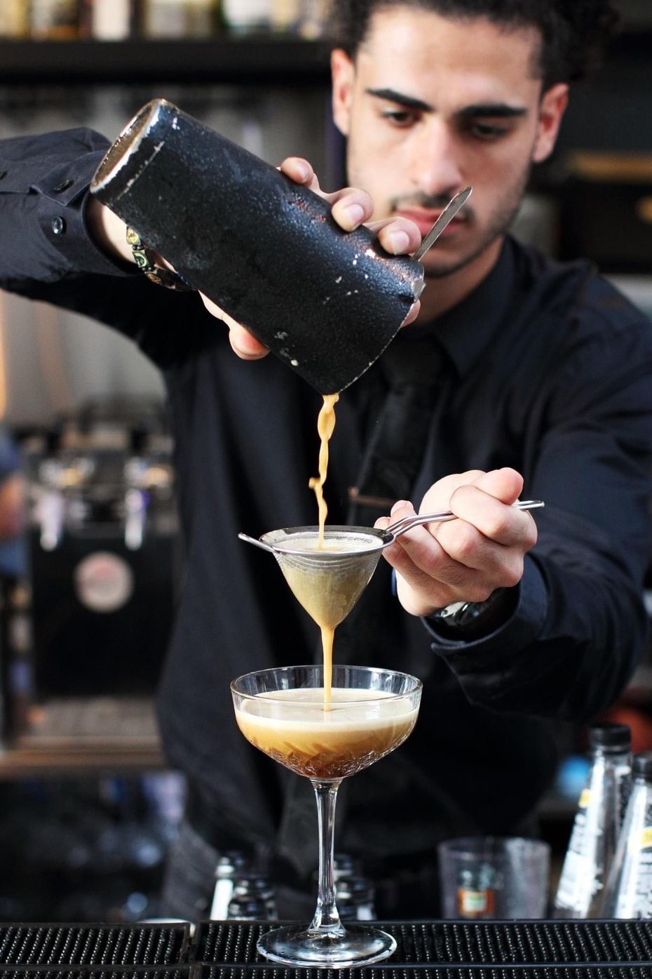 Bartender pouring a cocktail from a shaker through a strainer into a martini glass at a bar.
