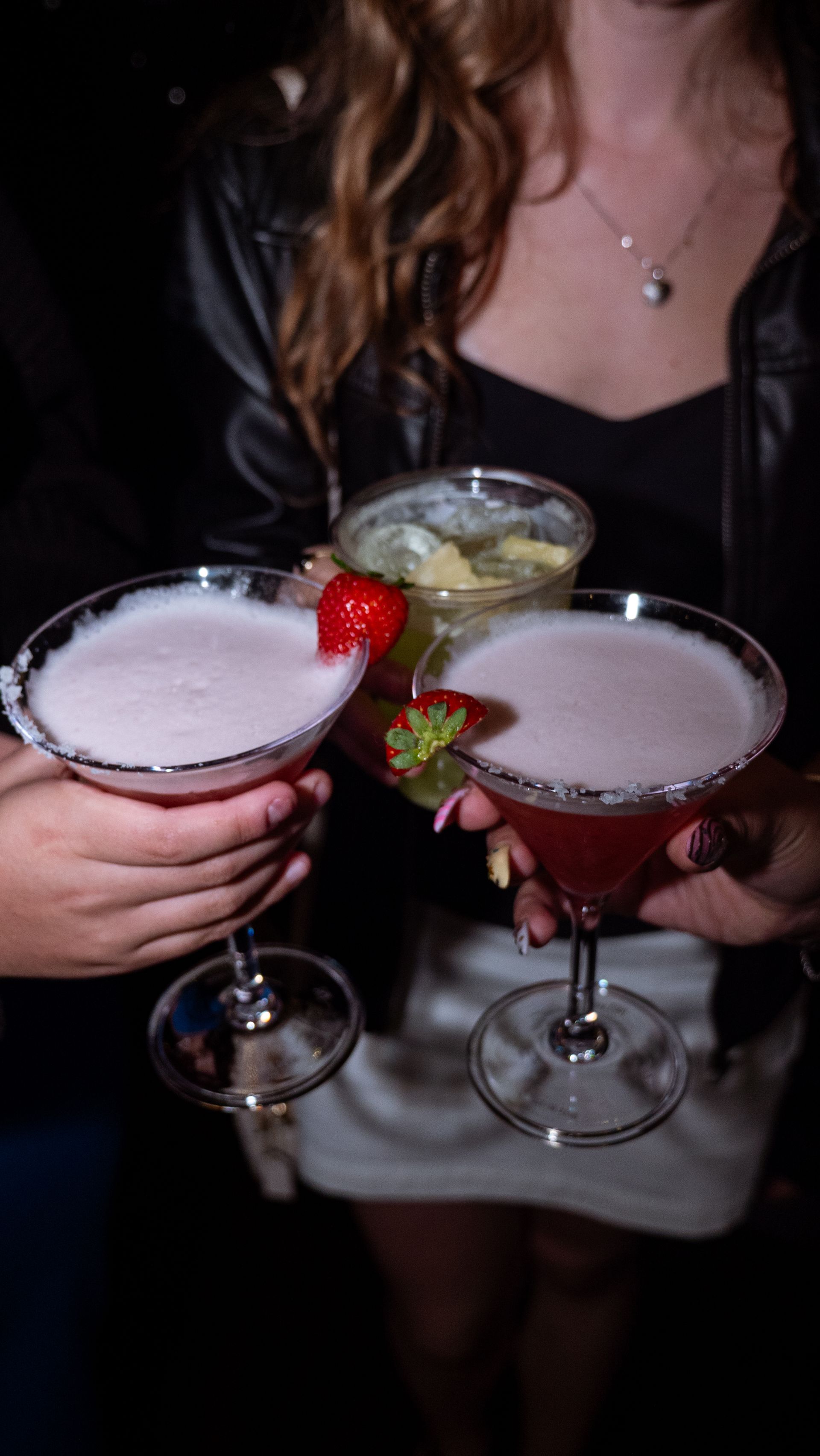 Hands holding three colorful cocktails with strawberry garnishes at a party.