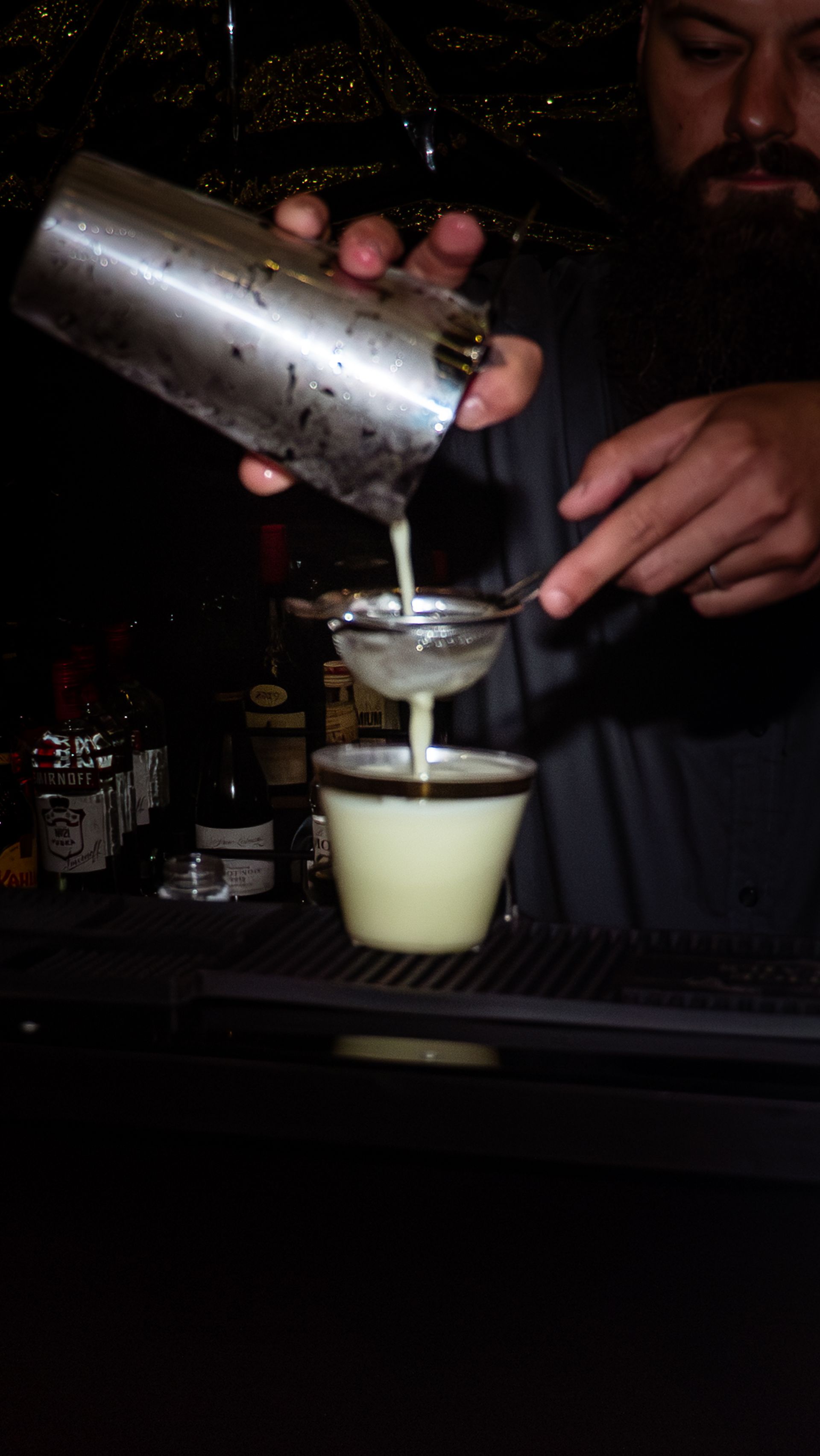 Bartender pouring a pale drink from a shaker through a strainer into a glass at a dim bar.