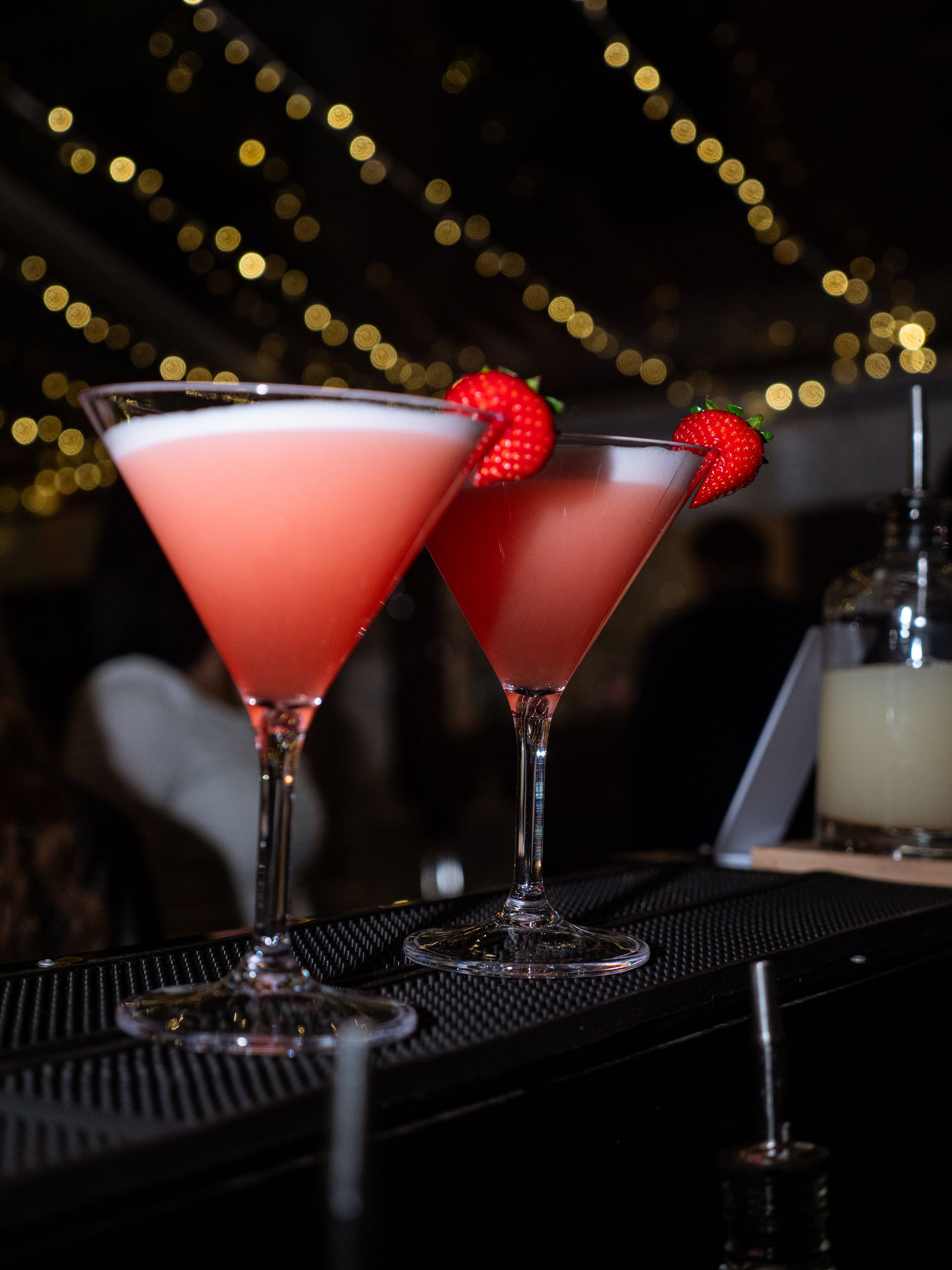Two red cocktails with raspberries on a bar tray, with soft bokeh lights in the background.