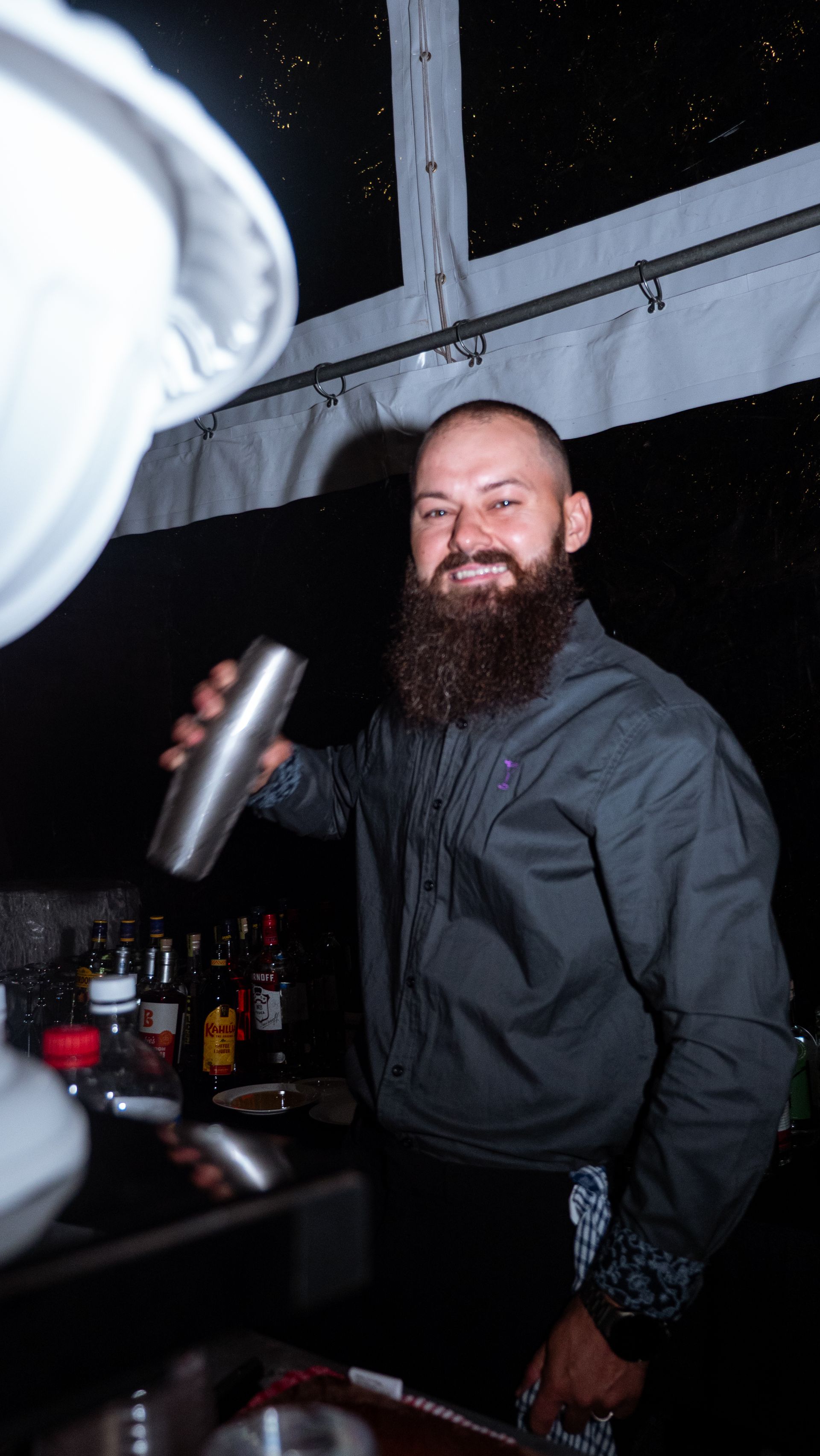 Bearded man in a dark shirt holding a metal can at a dimly lit bar table, smiling at the camera.
