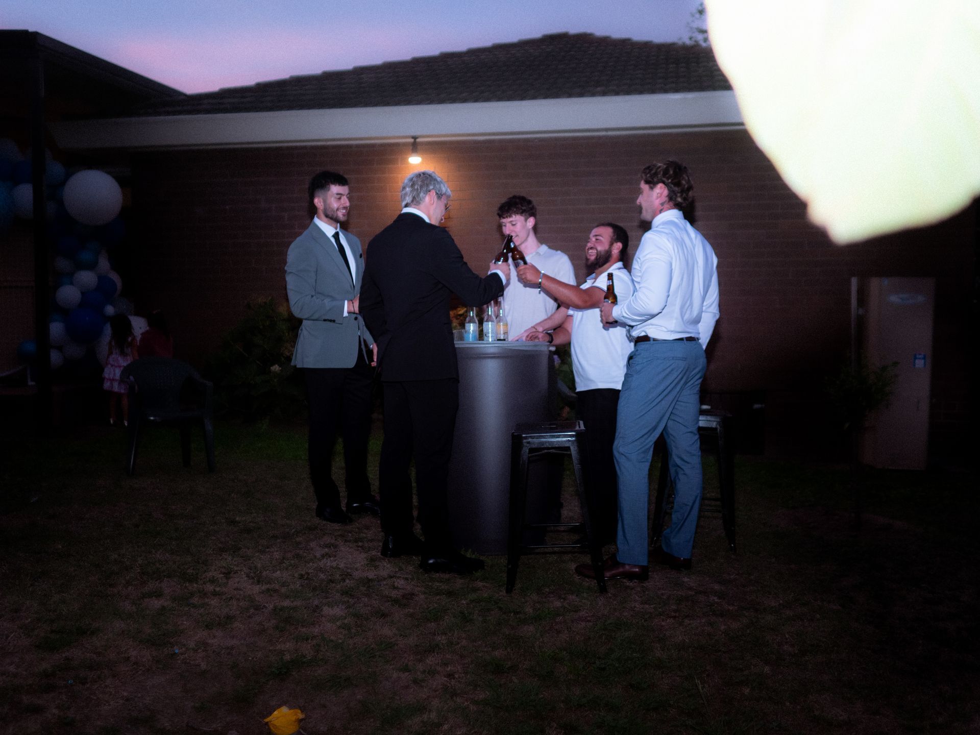 People gathered around a table under a tent at dusk, talking and holding drinks.