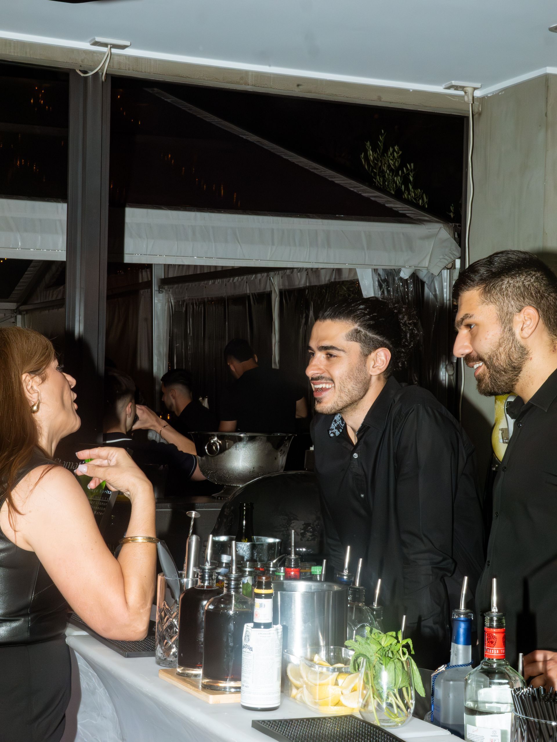 People chatting at a bar, with bottles on the counter and a dark, lively indoor setting.
