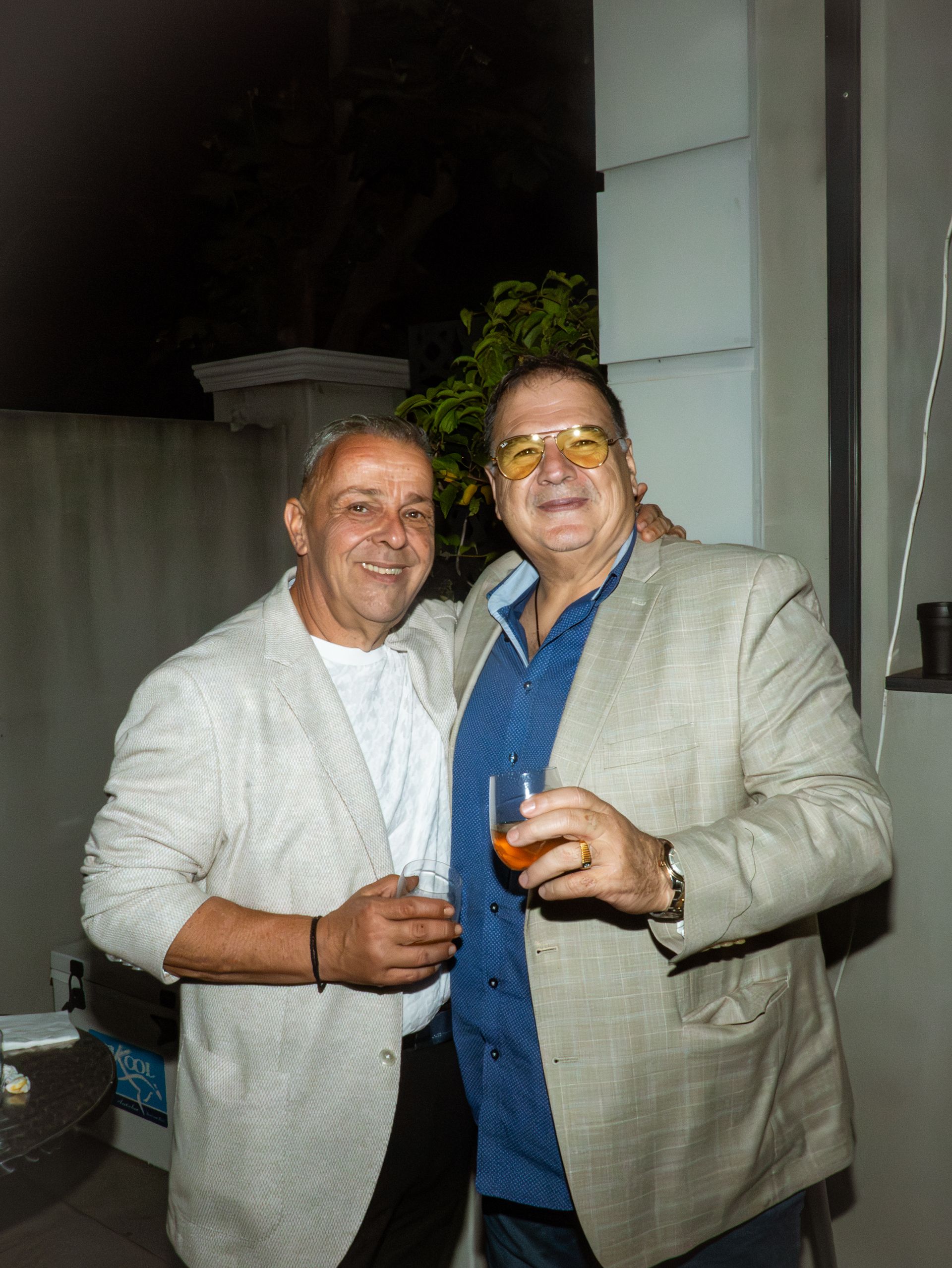 Two men in light jackets smiling and holding drinks at an evening gathering outdoors.