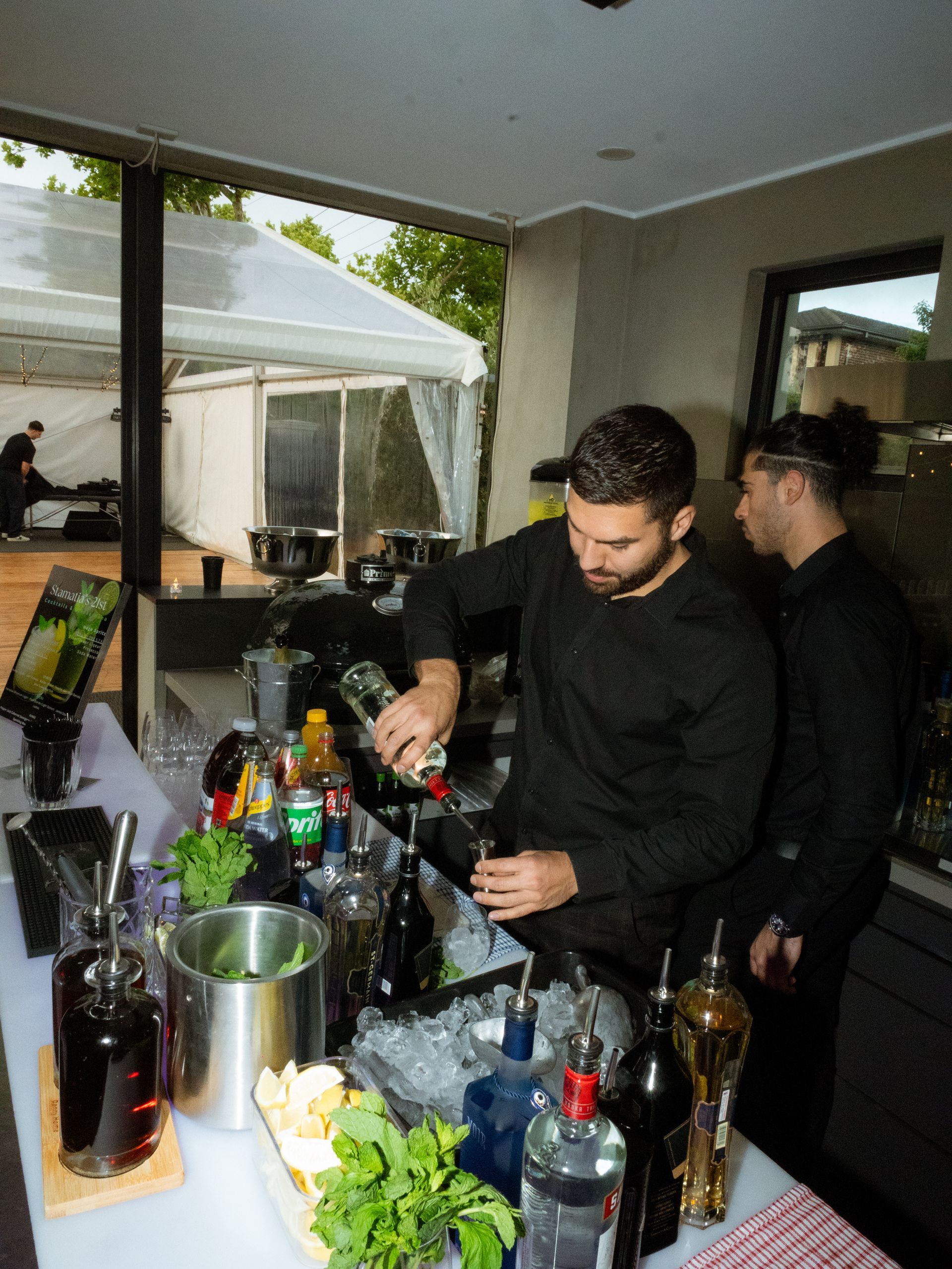Man mixing drinks at a bar counter with bottles, glassware, and a large window behind him